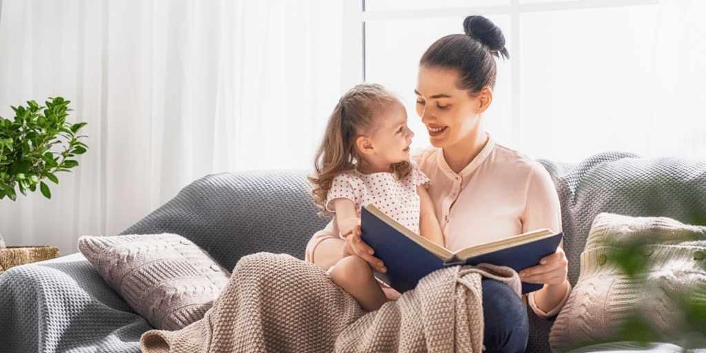 A smiling mother and young daughter reading a book together on a couch, representing families navigating North Carolina child support guidelines.