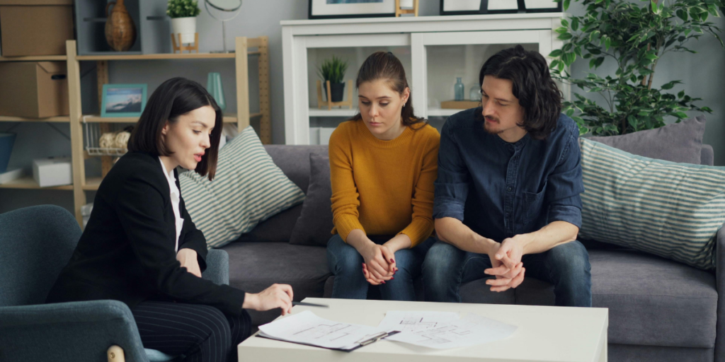 Woman in a black blazer reviewing documents on a coffee table with a young couple seated on a couch in a living room